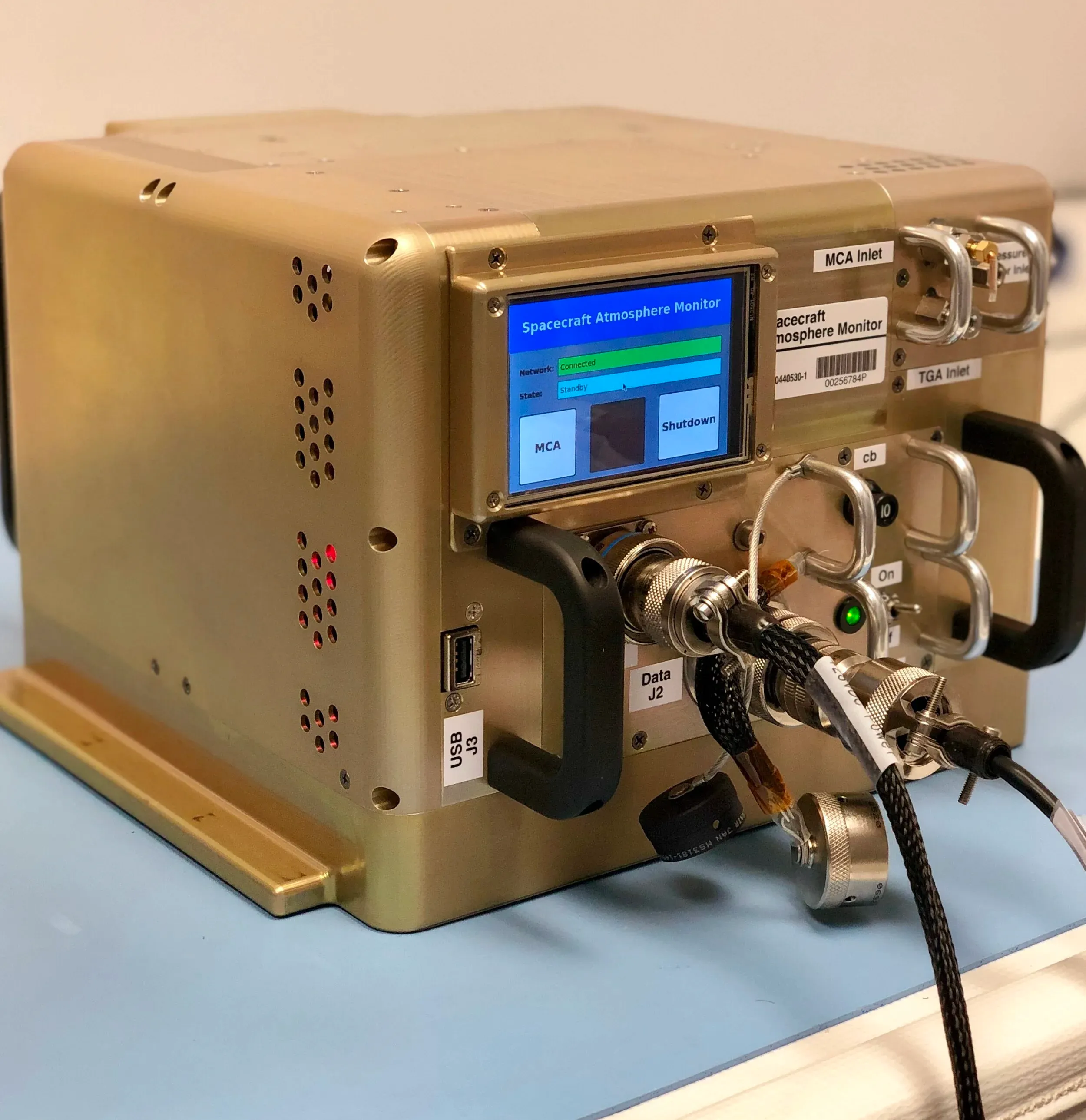 Gold-colored Spacecraft Atmosphere Monitor flight instrument with touchscreen and electrical connectors.